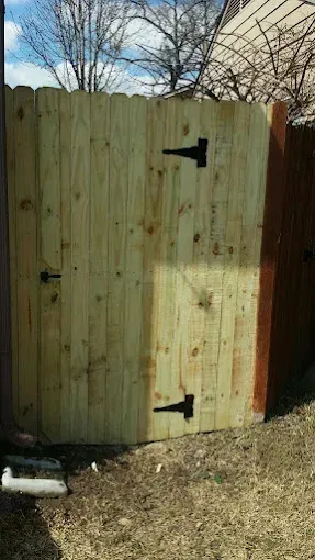 A new, light-colored wooden fence gate with black hinges installed against an older, dark wood fence.