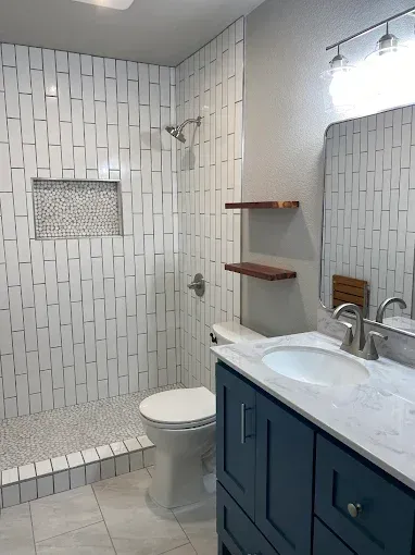 A modern bathroom featuring a blue vanity, white marble countertop, floating wood shelves, and a tiled walk-in shower.