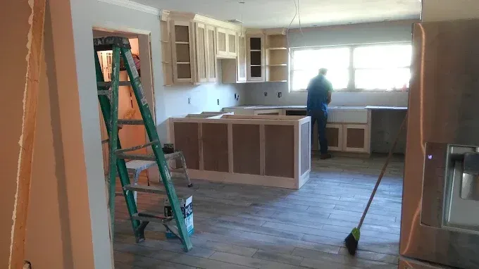 A person works in a kitchen during renovation, featuring light-colored cabinets, an island, and new wood-look flooring.