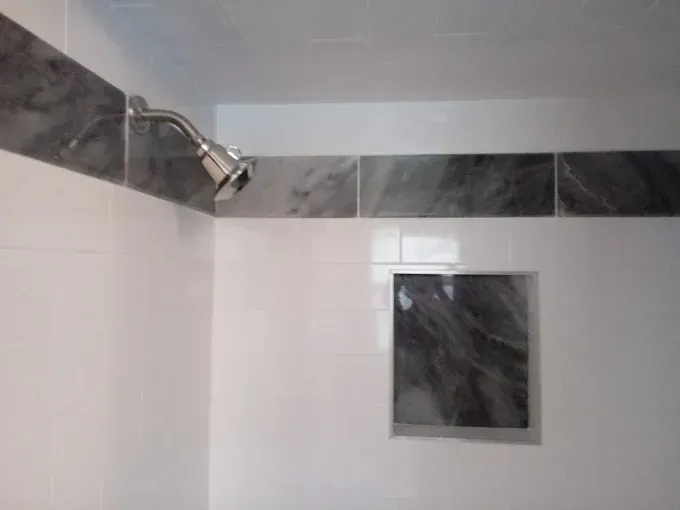 A shower stall with white wall tiles, a grey marble accent border, a matching marble soap niche, and a showerhead.