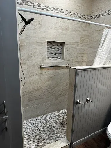 A walk-in shower with beige tile walls, a mosaic tiled floor and niche, a horizontal grab bar, and a side privacy wall.