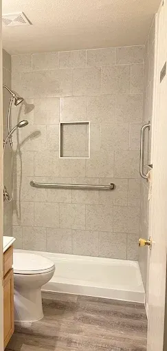 Accessible walk-in shower with light grey tiled walls, a horizontal grab bar, a shower niche, and a handheld showerhead.