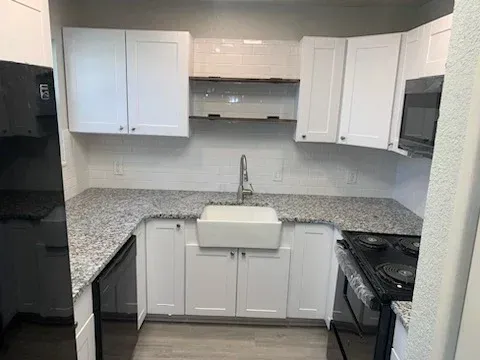 A modern kitchen featuring white cabinets, granite countertops, a farmhouse sink, and black appliances.
