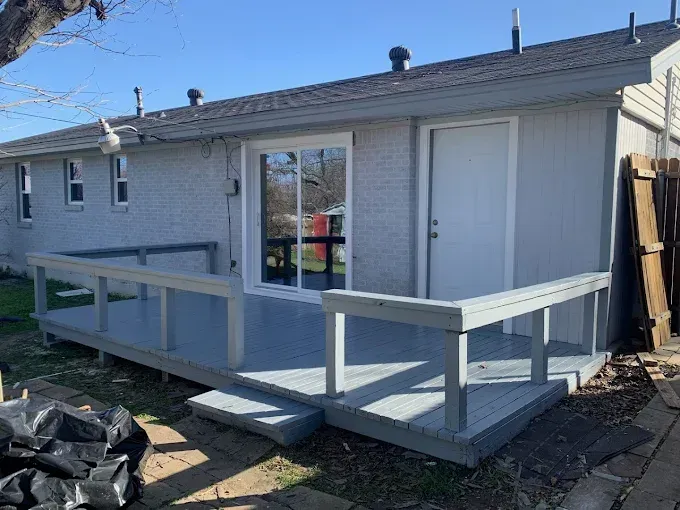 A light gray house exterior featuring a painted wood deck with railings, a sliding glass door, and a white side door.