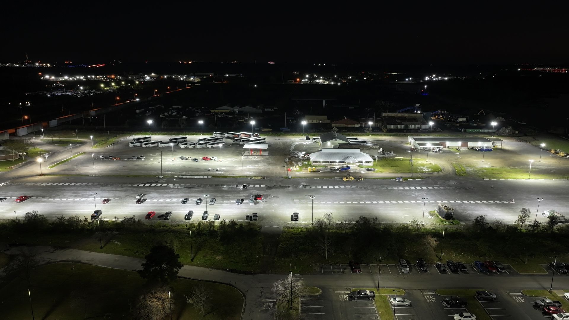 Aerial night view of a brightly lit parking lot with rows of cars and buildings in the distance.