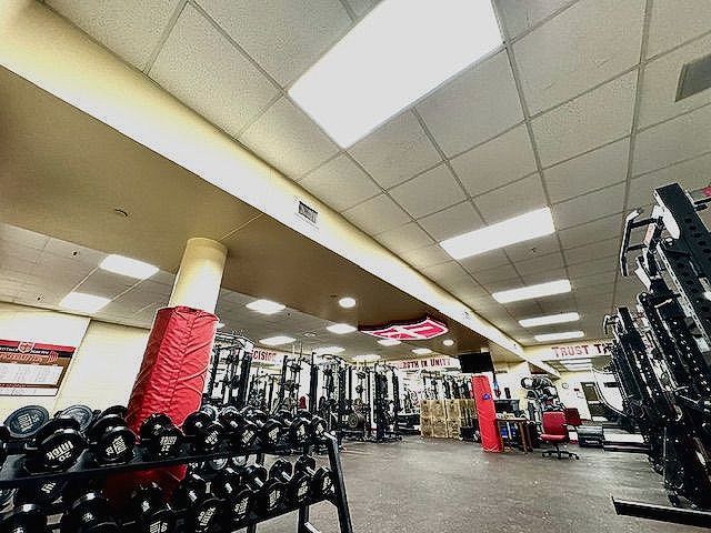 Interior of a gym with dumbbell racks, weight machines, and red columns under bright ceiling lights