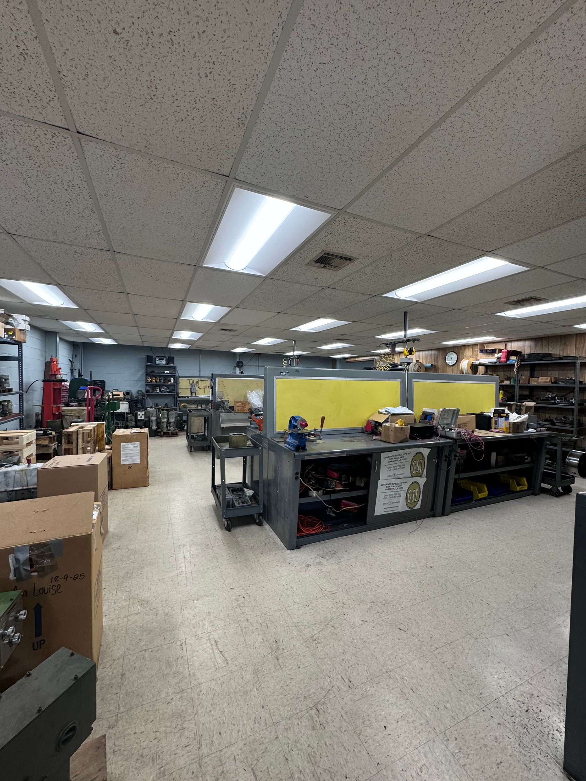 Cluttered workshop with workbenches, tools, boxes, and fluorescent ceiling lights under a drop ceiling.