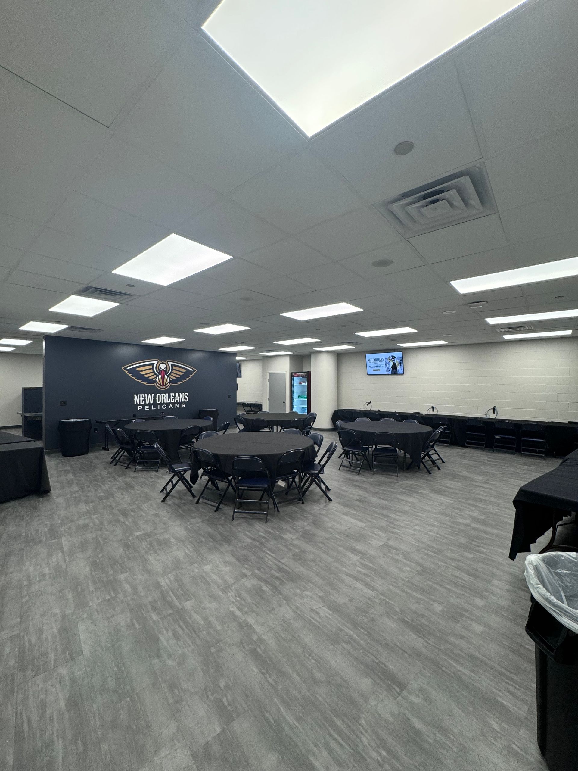 Empty conference room with tables and chairs, bright ceiling lights, and a wall-mounted screen.