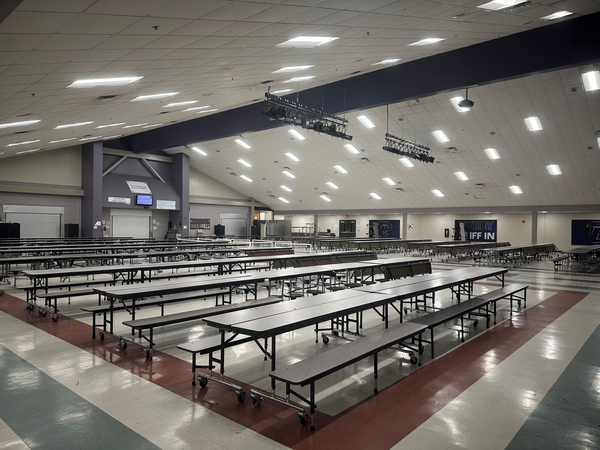Empty cafeteria with long rows of metal tables and benches under bright ceiling lights.