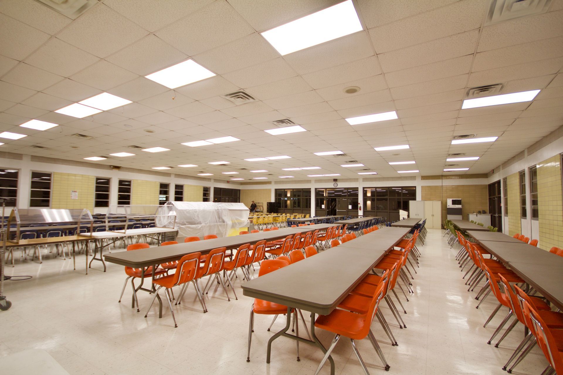 Empty cafeteria with long tables and rows of orange chairs under bright ceiling lights