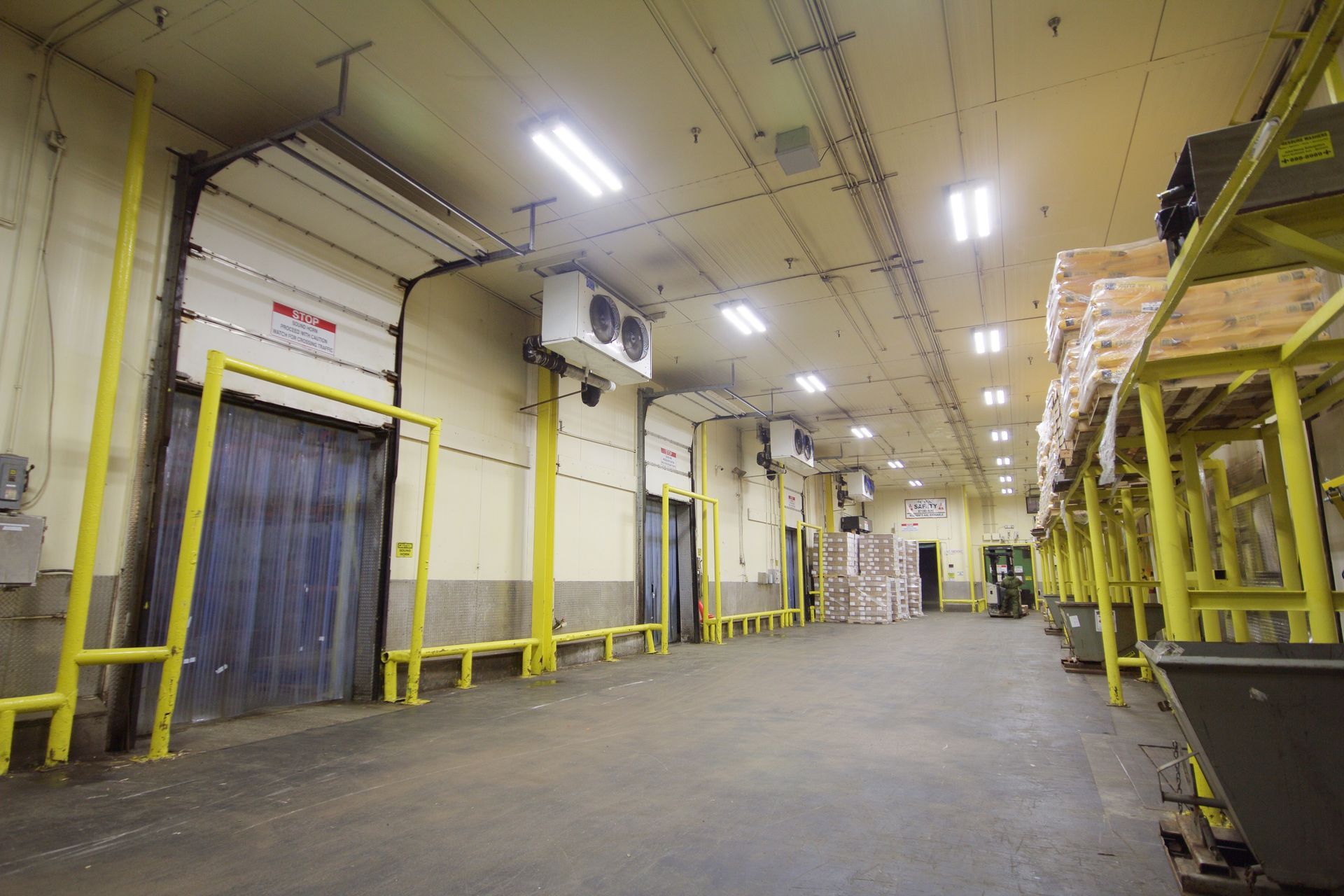 Empty warehouse aisle with yellow safety rails, concrete floor, and overhead lights.