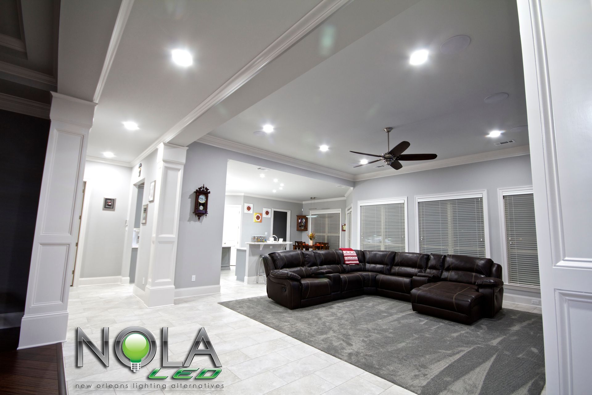 Spacious modern living room with dark sectional sofa, white walls, ceiling fan, and shuttered windows