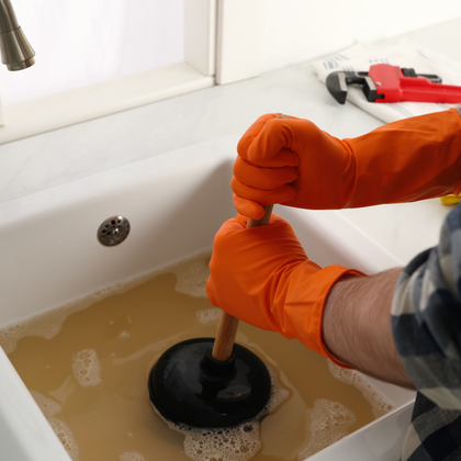 JM Plumbing Inc | A person wearing orange gloves is using a plunger to clear a sink