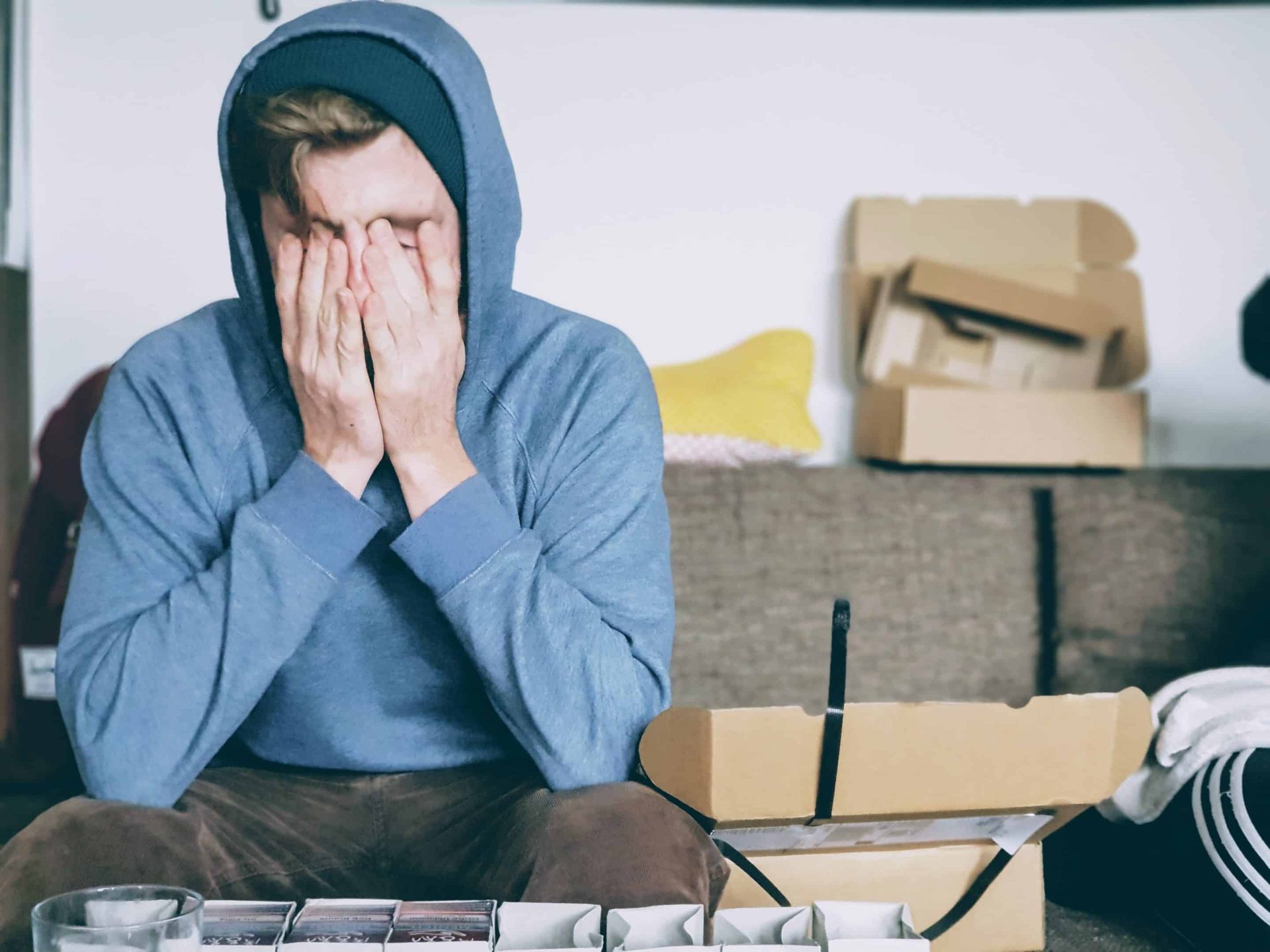 Man in blue hoodie, head in hands, appears stressed, seated on couch with cardboard boxes nearby.