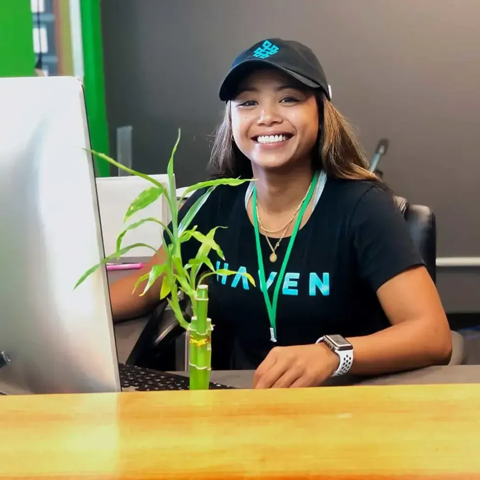 Woman smiling at a desk, wearing a hat and t-shirt with “Haven” on it; green bamboo plant on the desk.