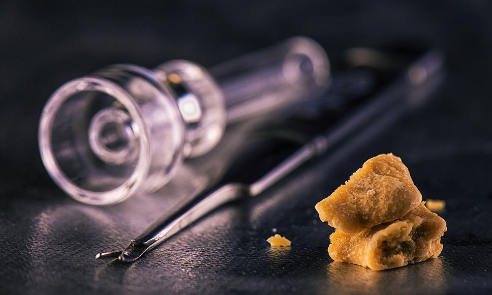 Dabs setup with glass rig, dab tool, and golden-brown concentrate on a dark surface.