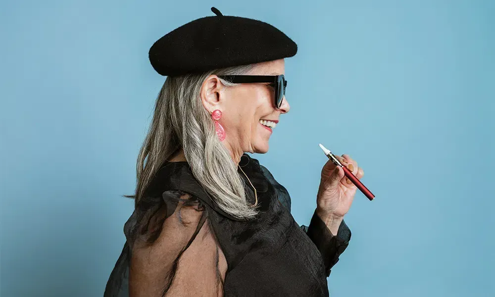 Woman in beret and sunglasses smiles, holding a pen. Against blue background.
