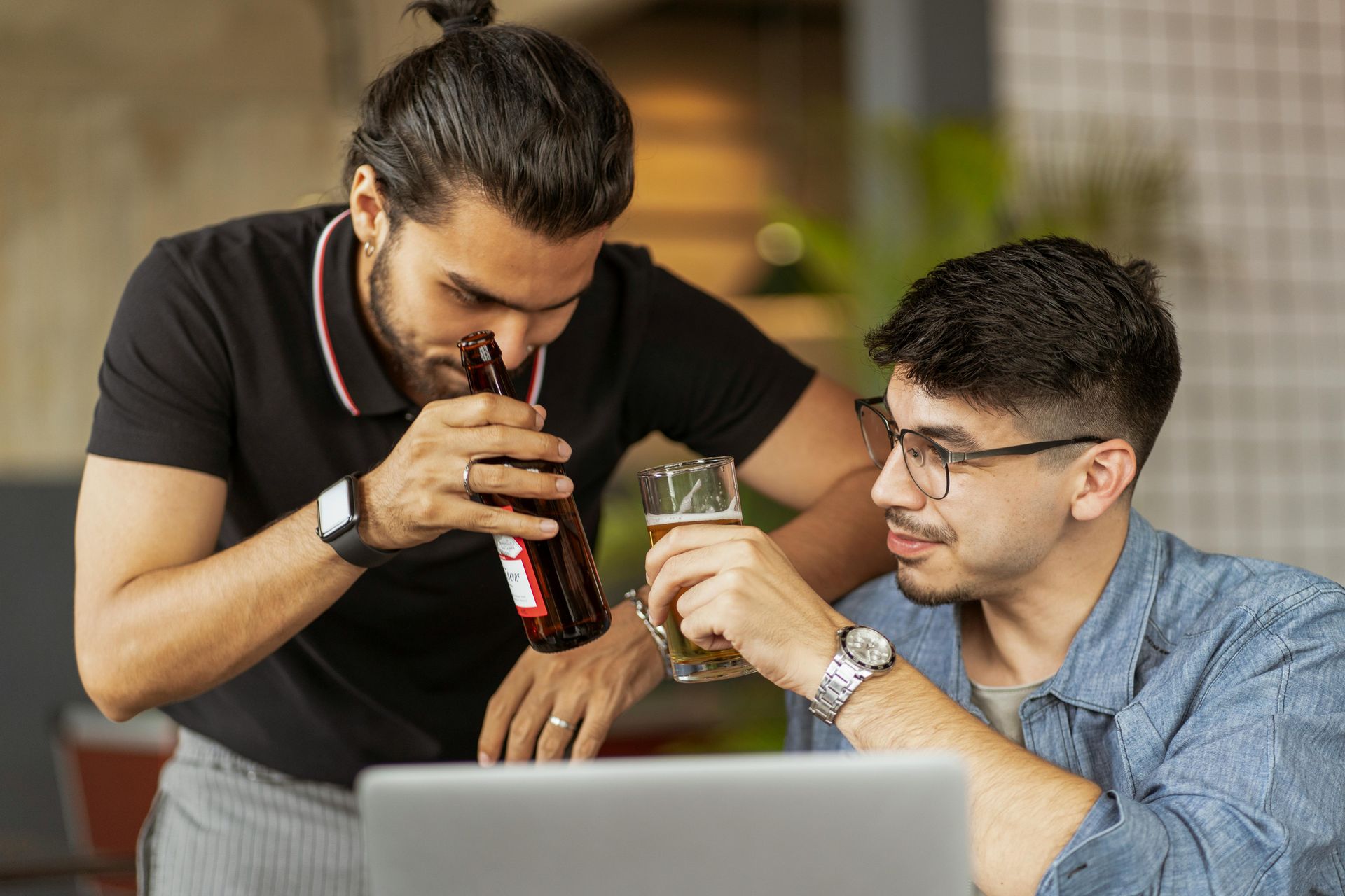Two men, one drinking from a bottle, the other holding a beer glass near a laptop. Indoors.