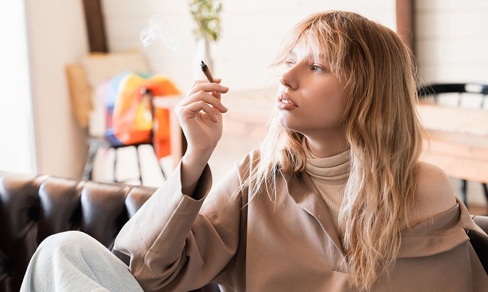 Woman holding a lit cannabis joint, exhaling smoke, in an indoor setting.