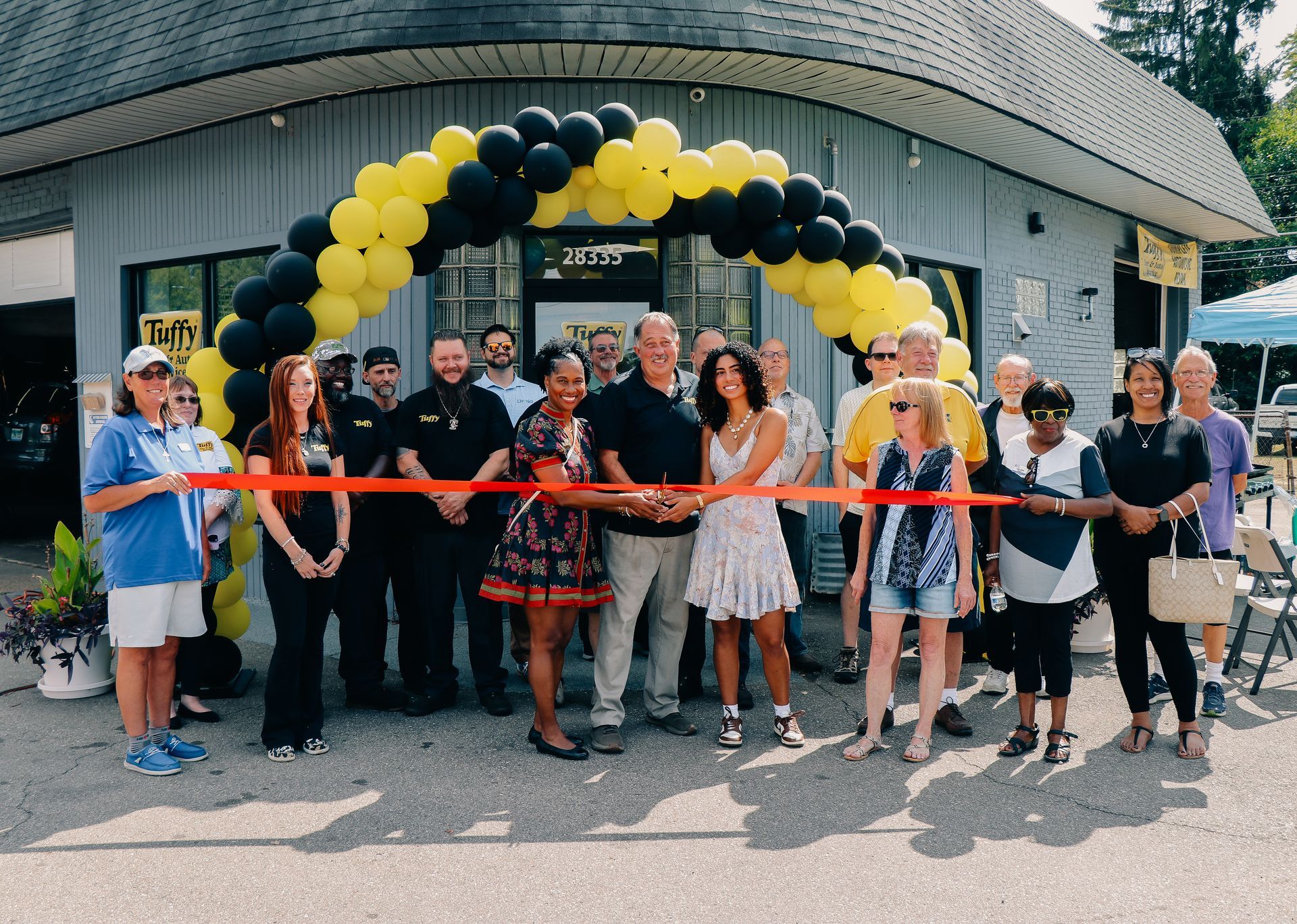 People cutting a red ribbon at a grand opening under yellow and black balloon arch.