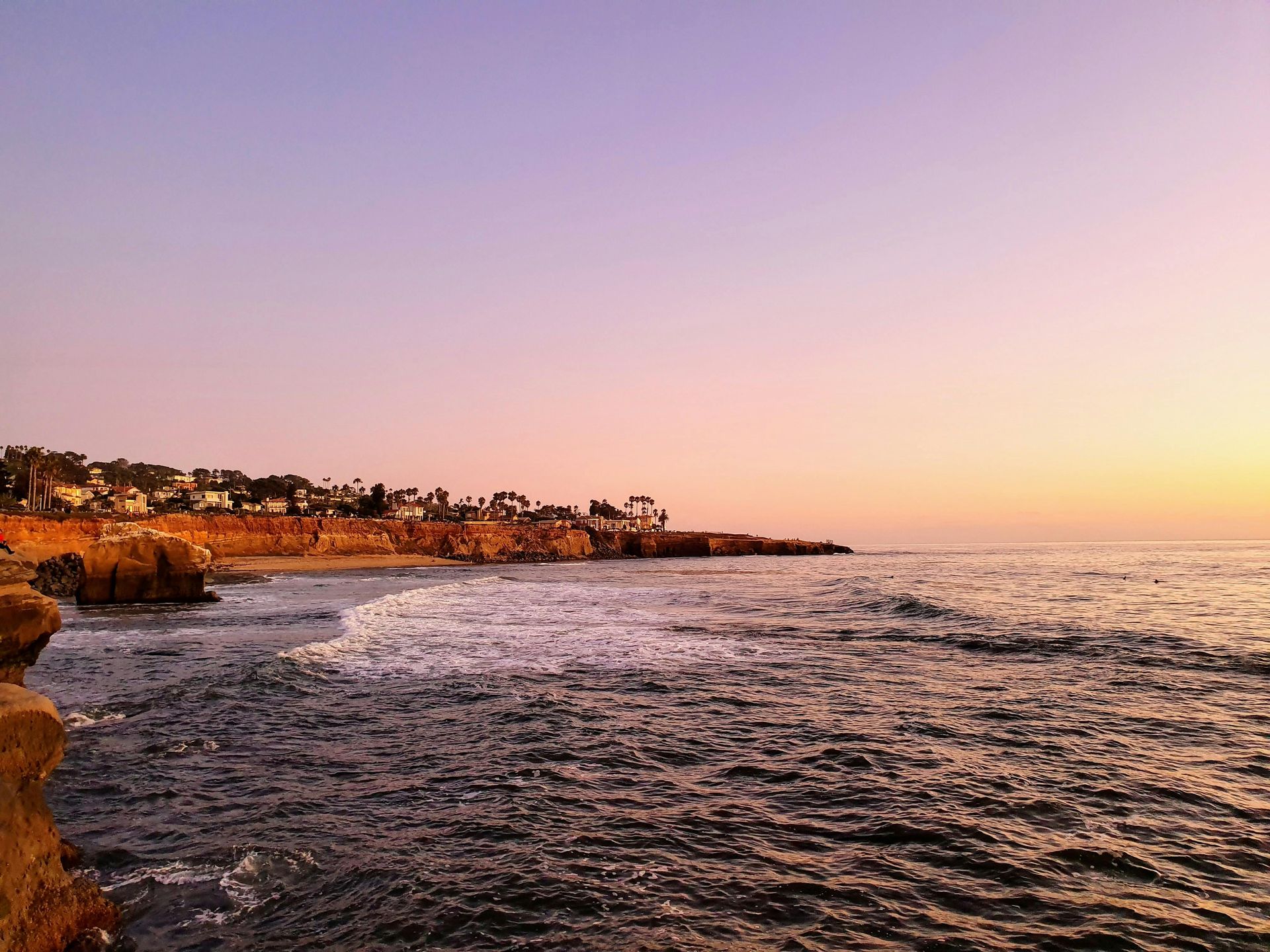 Ocean at sunset with a rocky coastline, pink and purple sky, calm water.