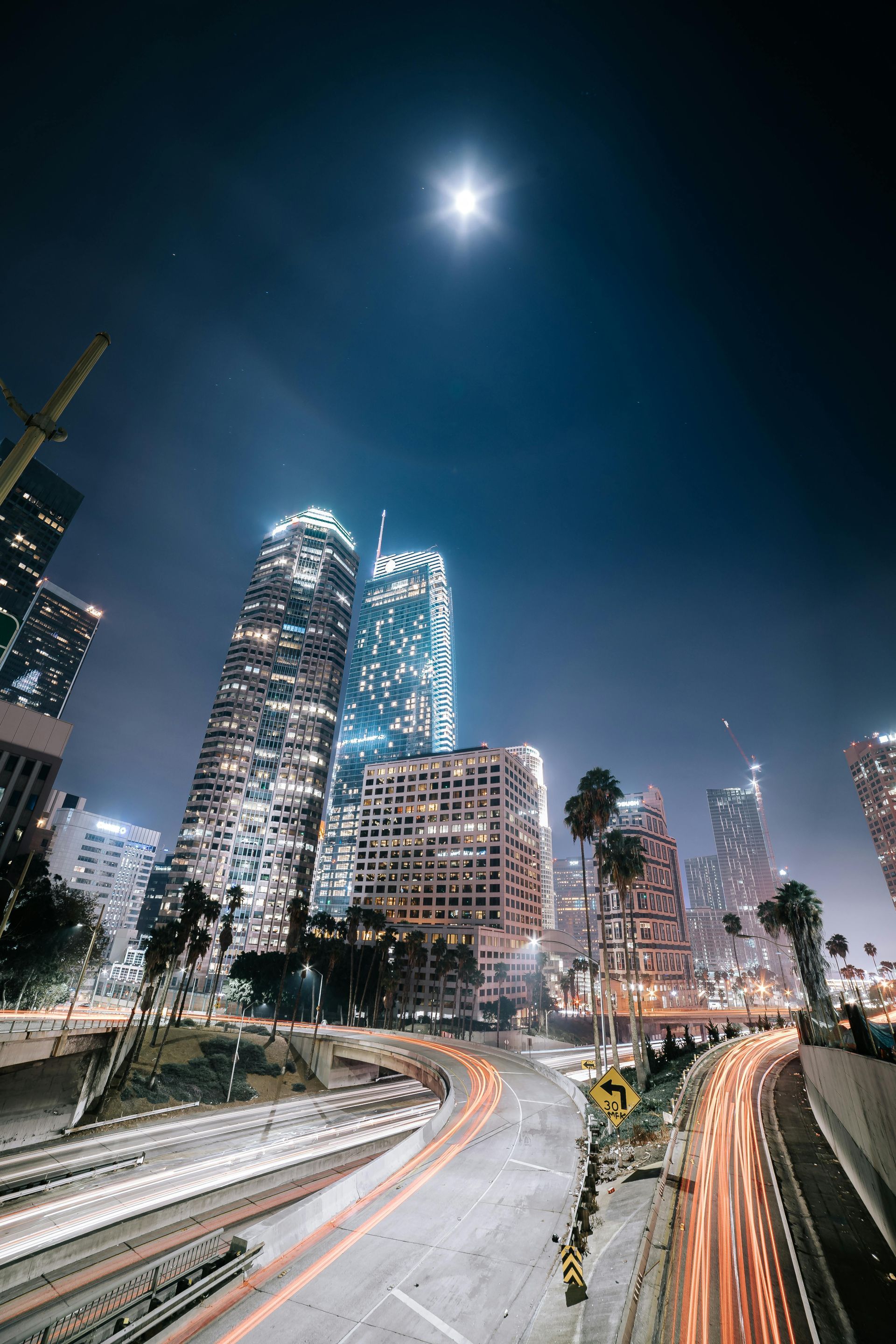 Nighttime cityscape with illuminated skyscrapers under a full moon. Traffic streaks along a curving road.