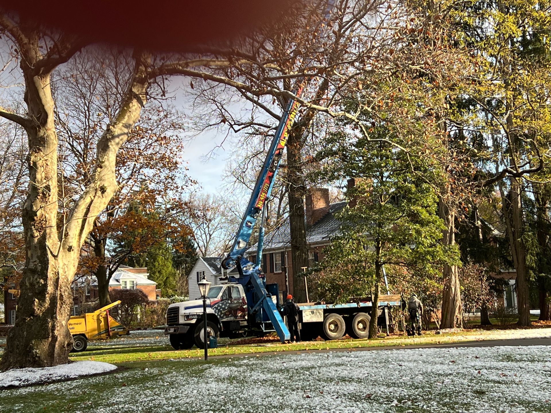 A crane truck with an extended boom performs tree trimming work in a snowy residential yard.