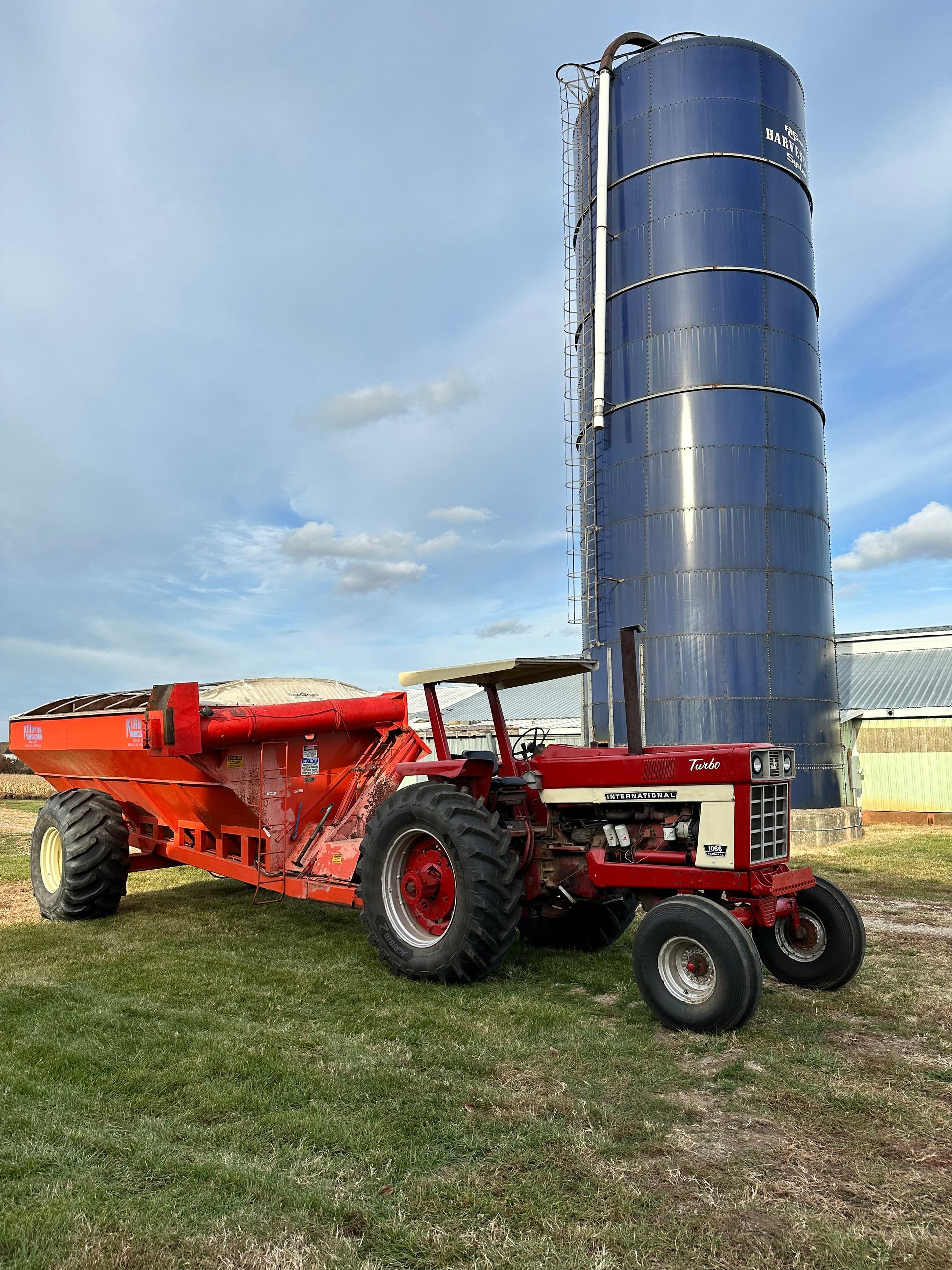 Red tractor pulling an orange grain cart next to a tall blue silo on a grassy field under a cloudy sky.