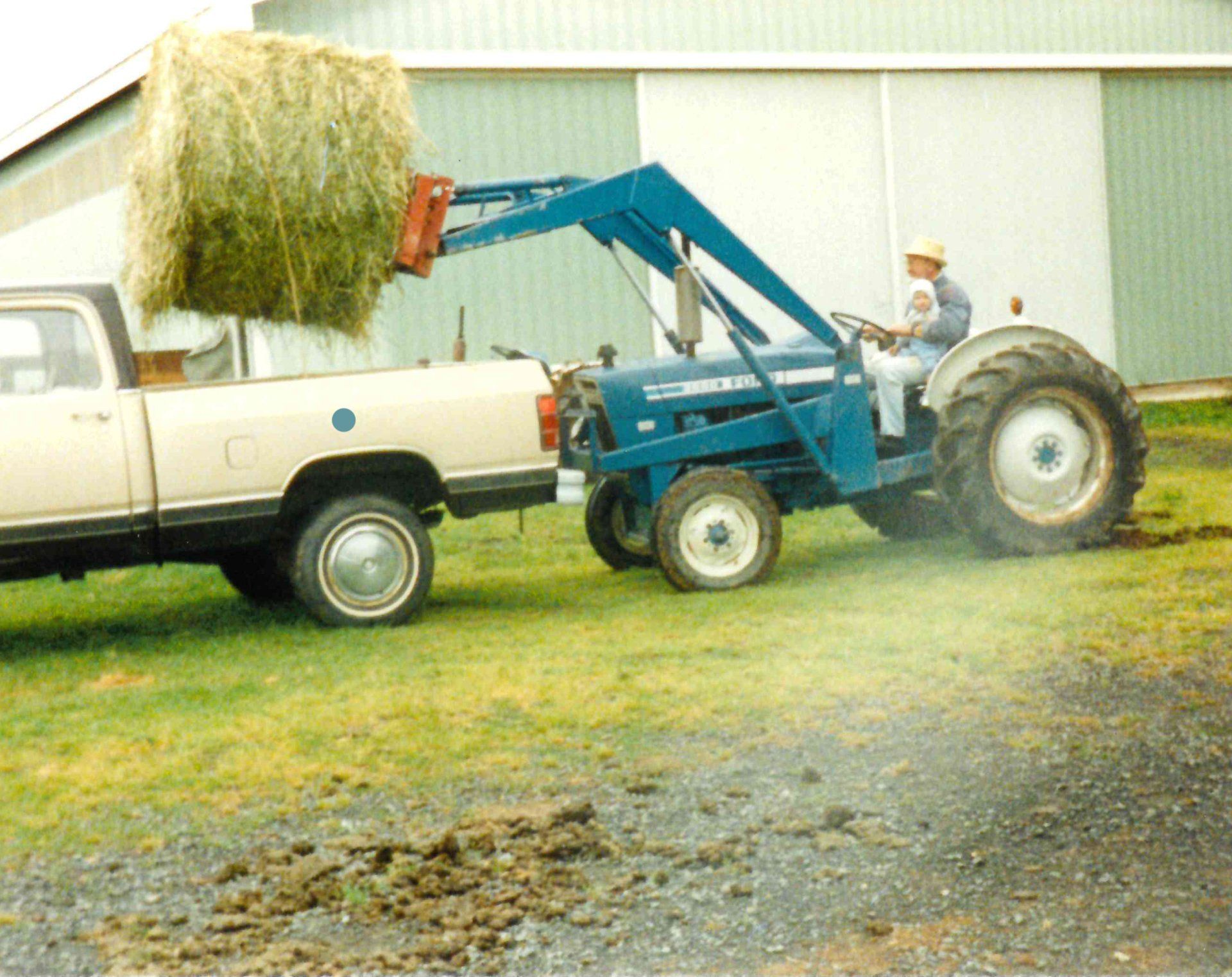 A tractor with hay bale lifting into a truck, in front of a white barn. A man drives the tractor.