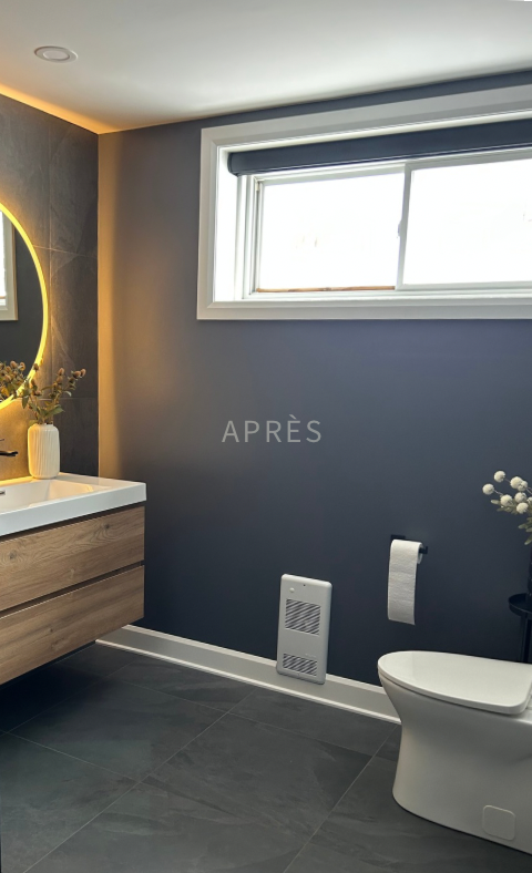 Salle de bains moderne avec murs sombres, miroir rond, meuble-lavabo en bois, toilettes blanches et petite fenêtre.