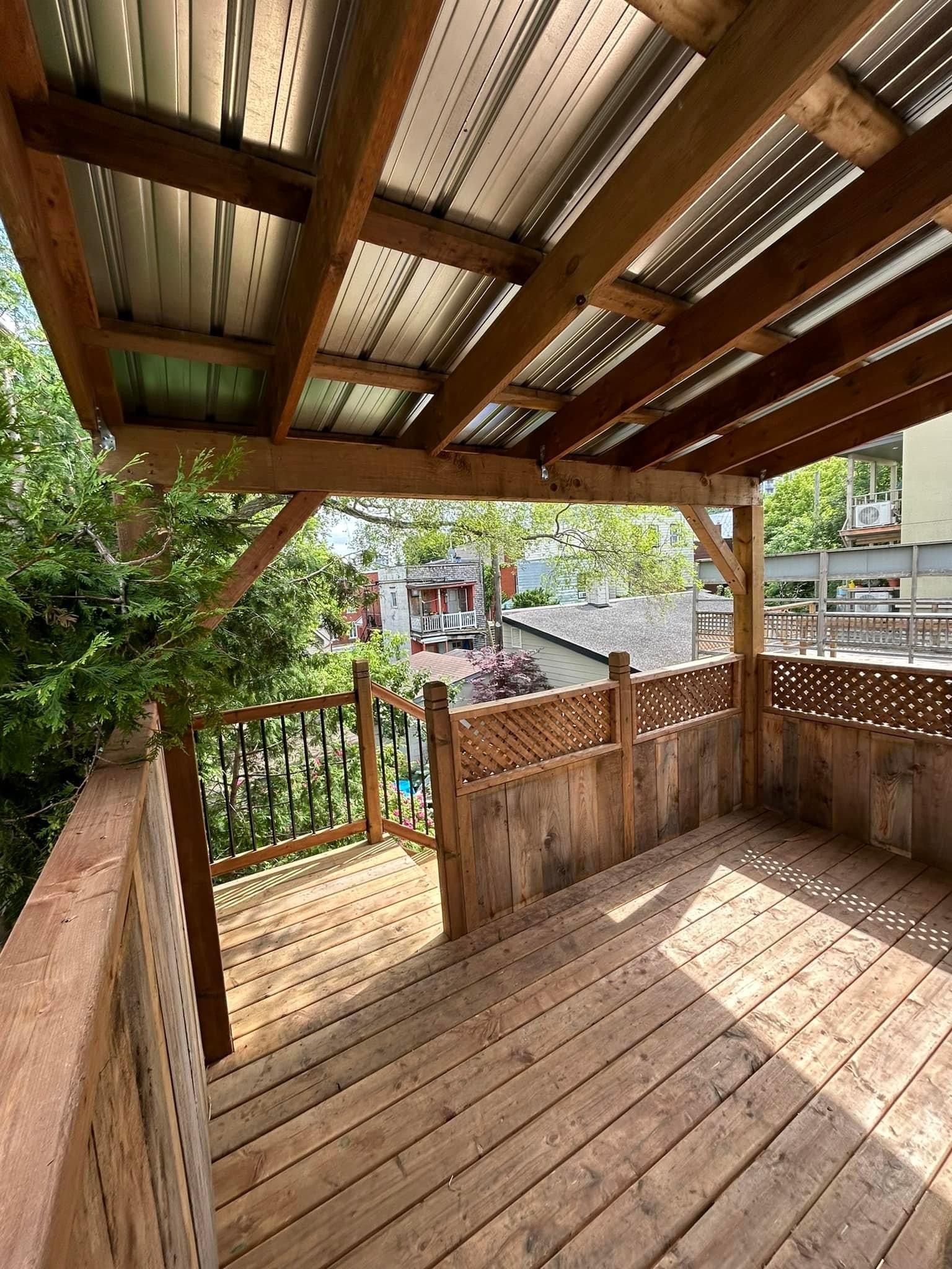 Terrasse en bois à l'arrière, sous un toit à claire-voie, donnant sur des arbres verdoyants et une maison voisine.