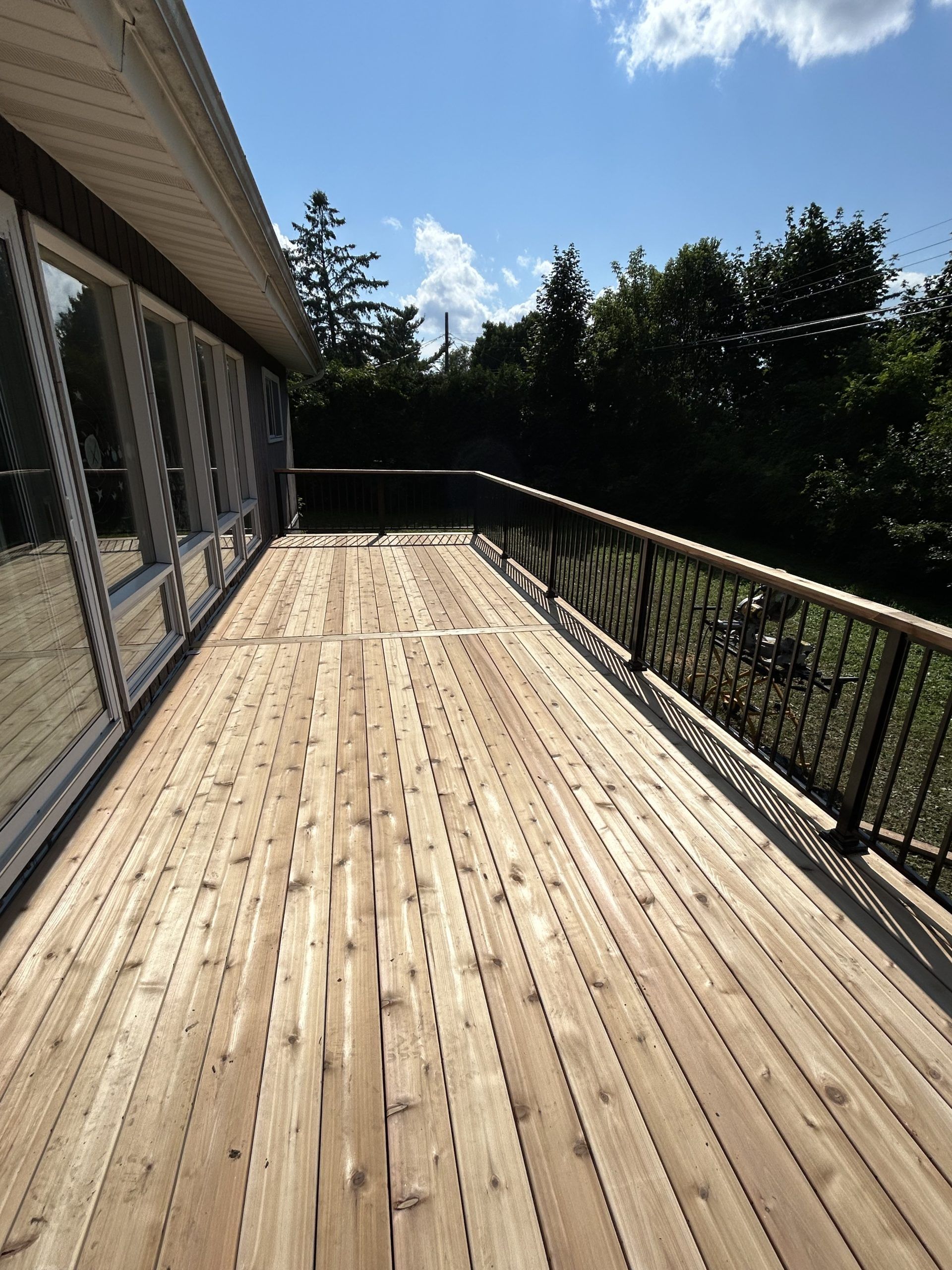 Terrasse en bois jouxtant une maison aux balustrades noires, bordée d'arbres sous un ciel d'un bleu éclatant.