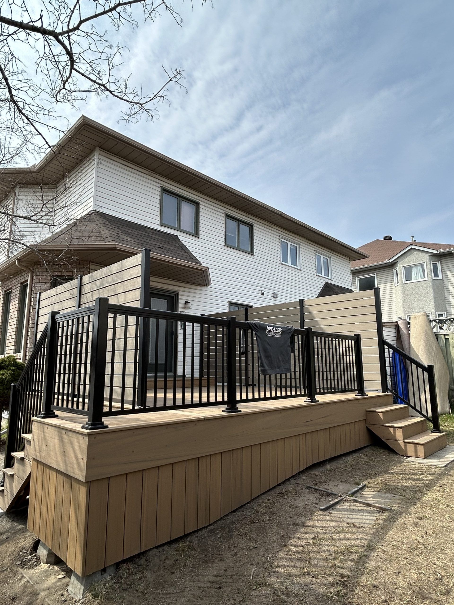Terrasse arrière avec balustrades noires jouxtant une maison blanche à deux étages sur un terrain en pente.