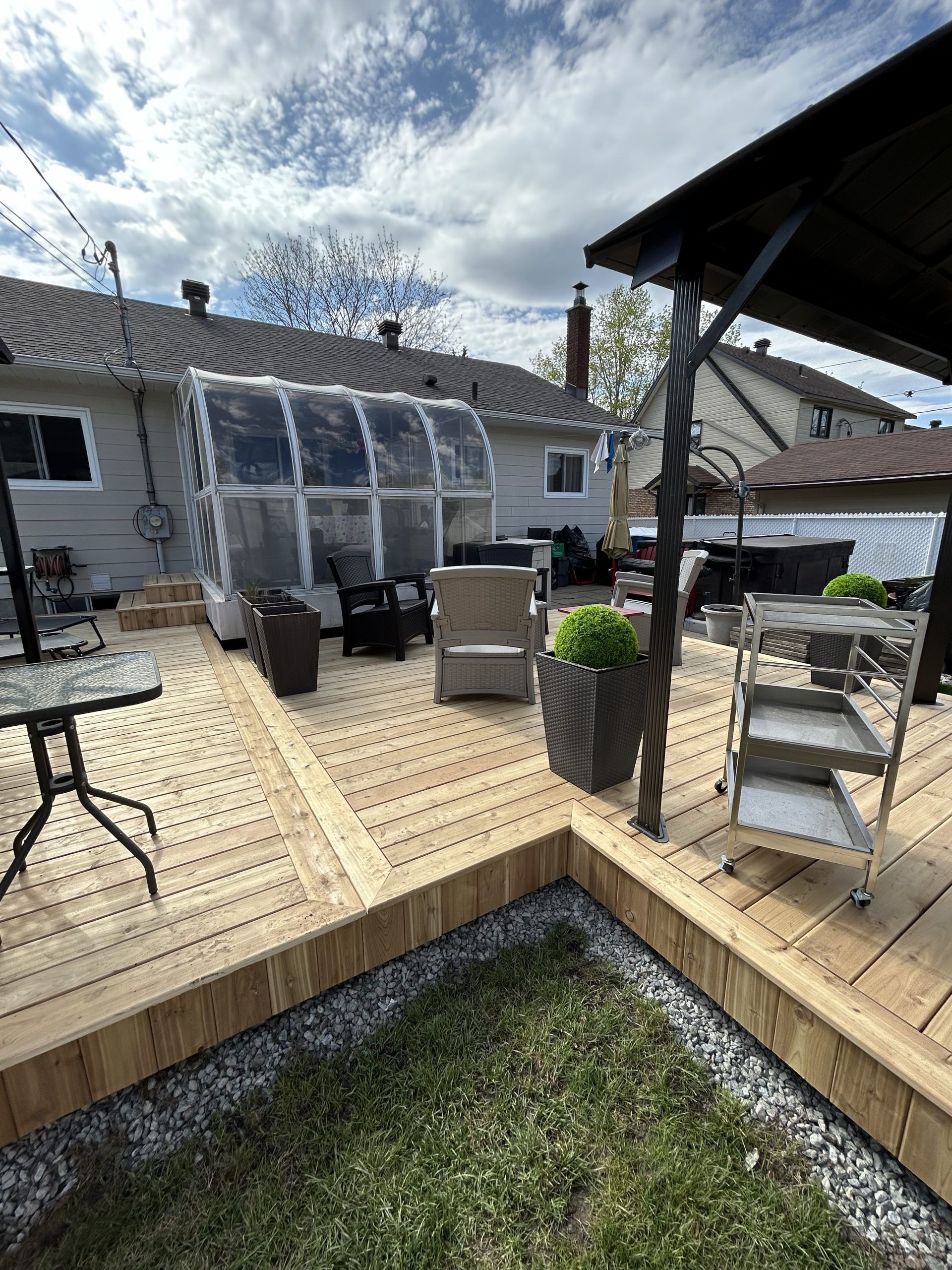 Terrasse en bois dans le jardin avec mobilier de jardin, plantes en pot et véranda sous un ciel nuageux
