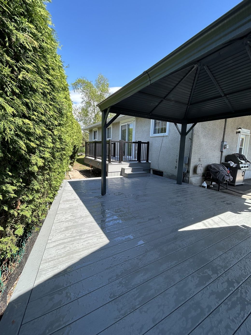 Terrasse en bois couverte jouxtant un bâtiment en pierre, bordée d'une haie verte.