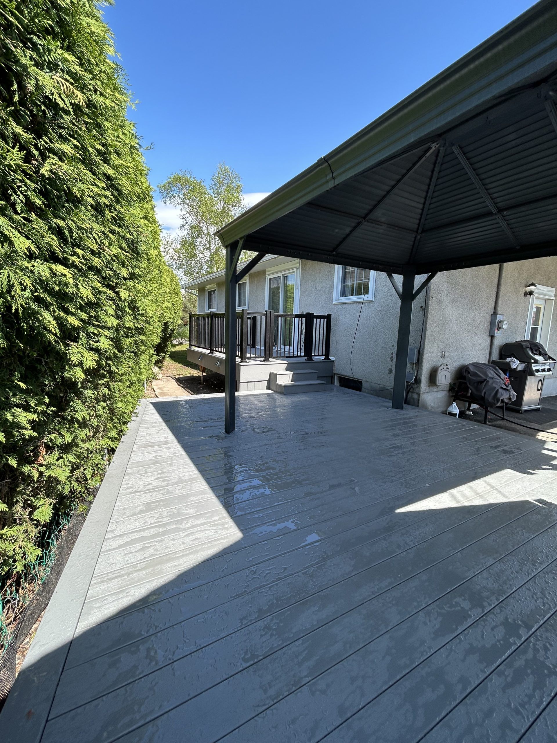 Terrasse en bois couverte jouxtant un bâtiment en pierre, bordée d'une haie verte.