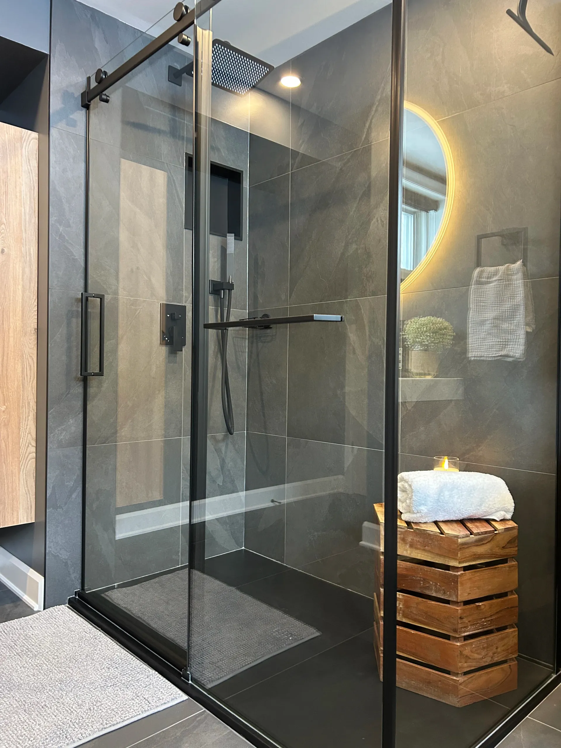 Douche à l'italienne en verre avec robinetterie noire, carrelage gris et banc en bois avec serviettes.