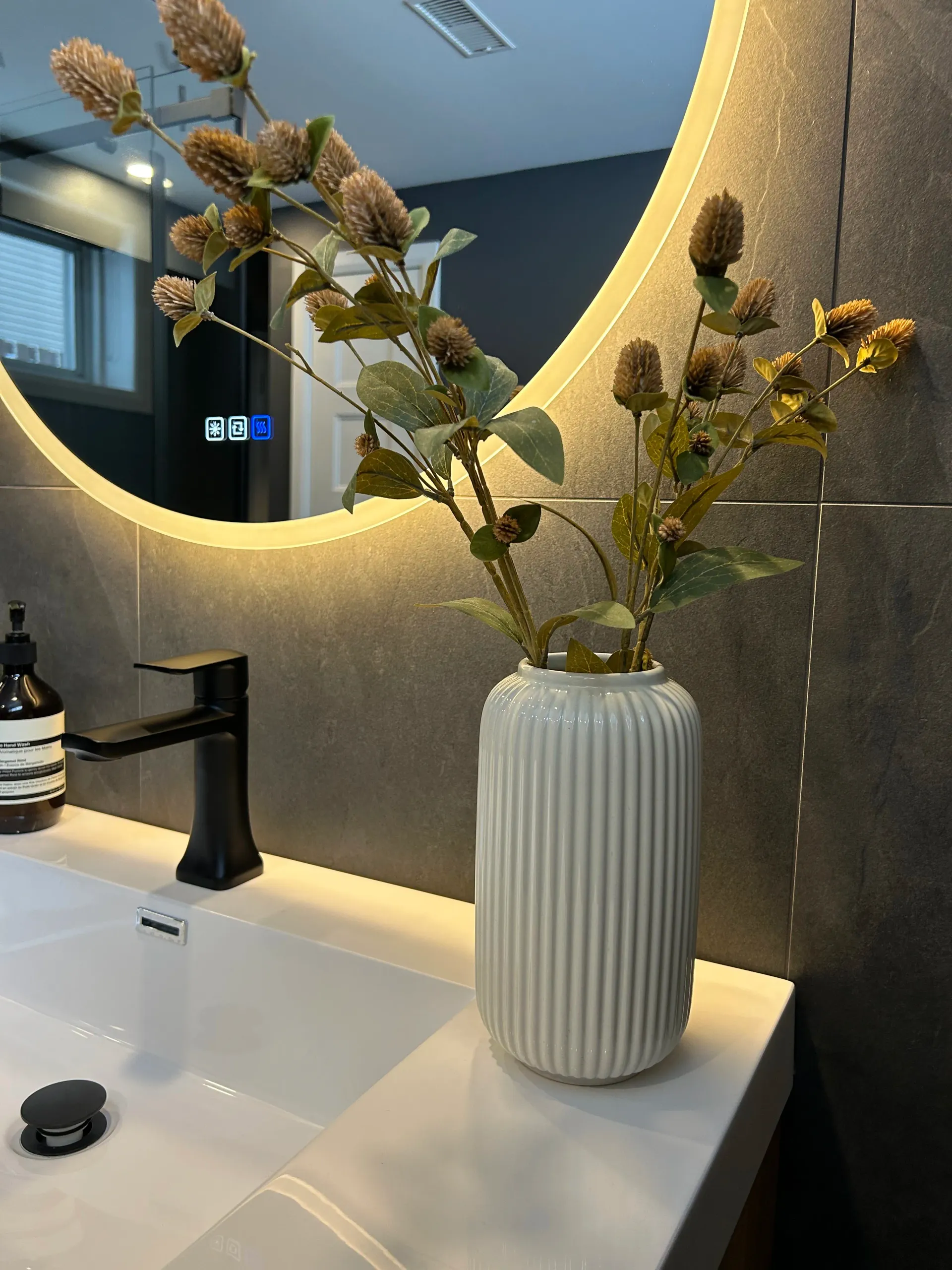 Vase de fleurs séchées sur un meuble-lavabo, à côté d'un miroir rond et d'un robinet.