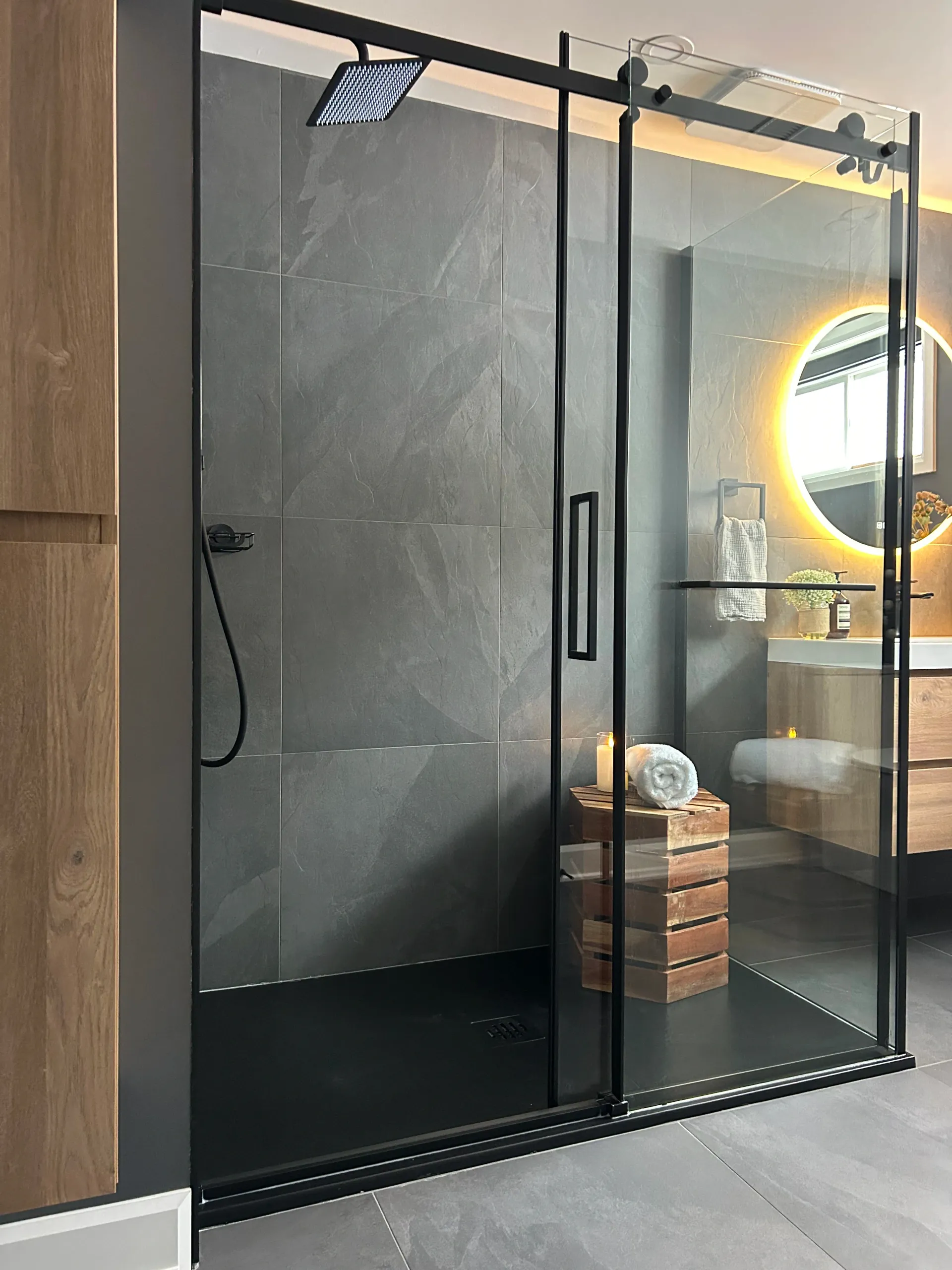 Douche de salle de bain moderne avec portes en verre à cadre noir et miroir rond éclairé en arrière-plan