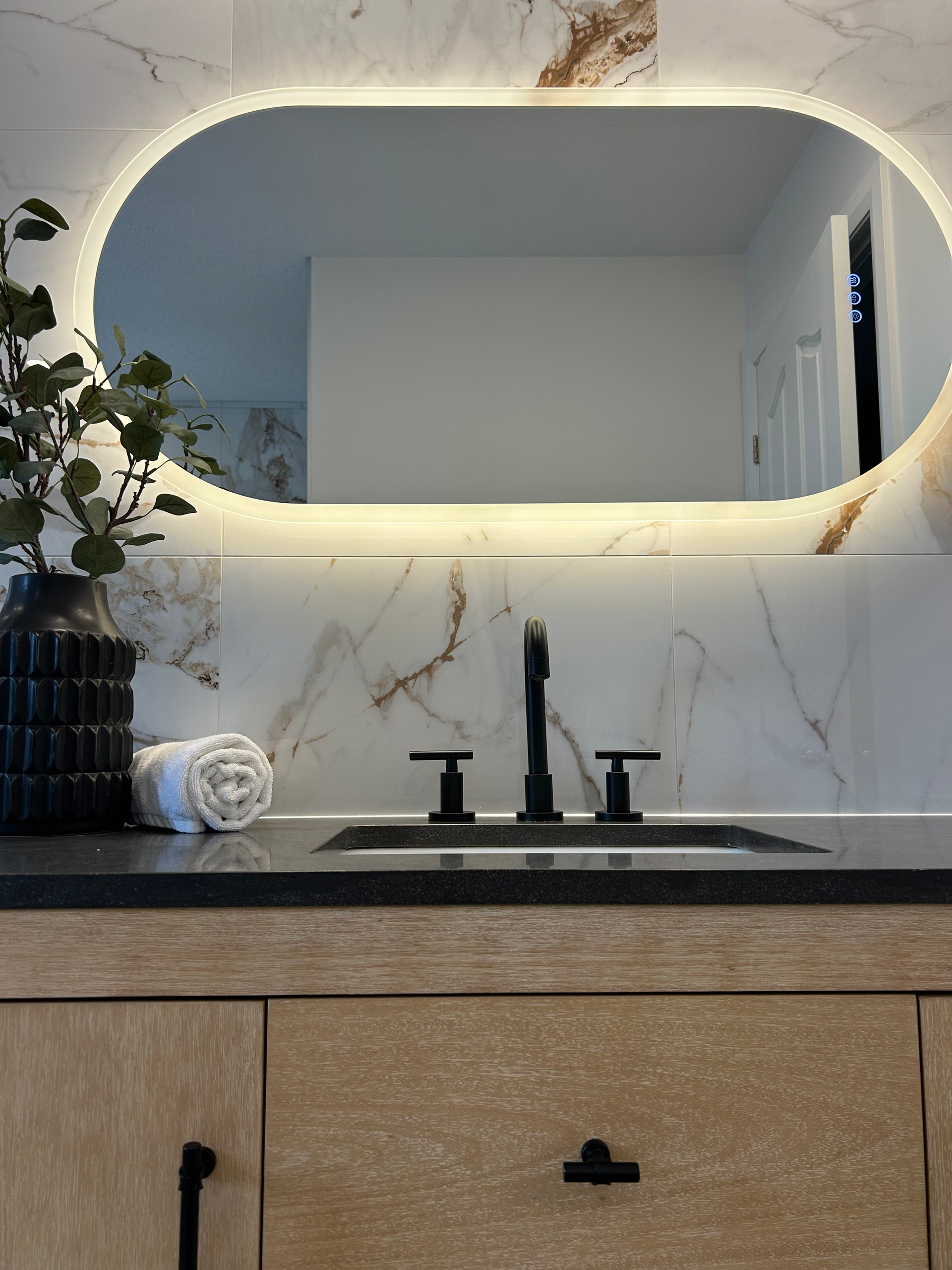 Meuble-lavabo moderne avec miroir ovale rétroéclairé, robinetterie noire, mur en marbre et armoire en bois.