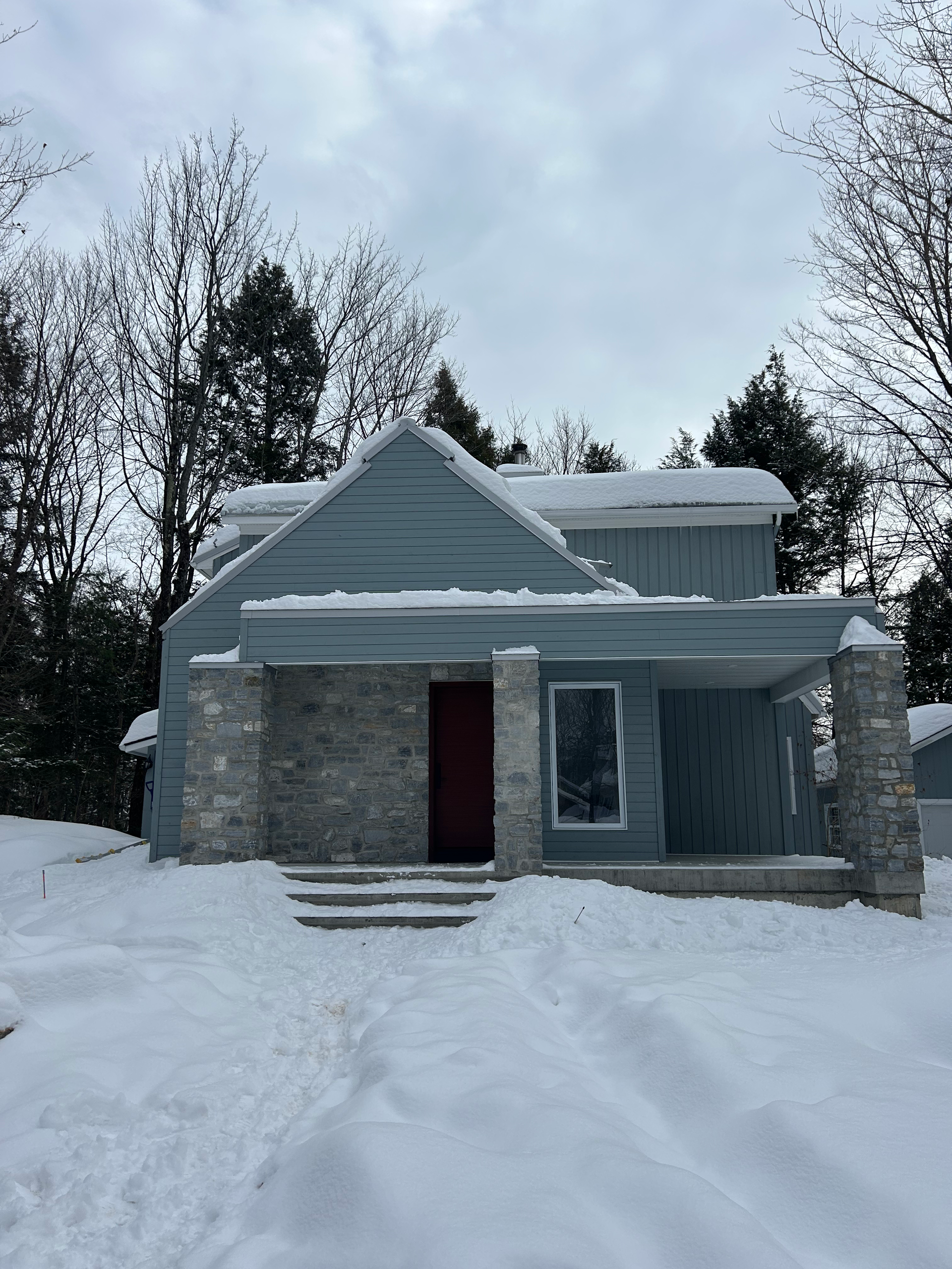 Maison en pierre enneigée, avec une porte et un porche rouge foncé, encadrée par des arbres dénudés en hiver.