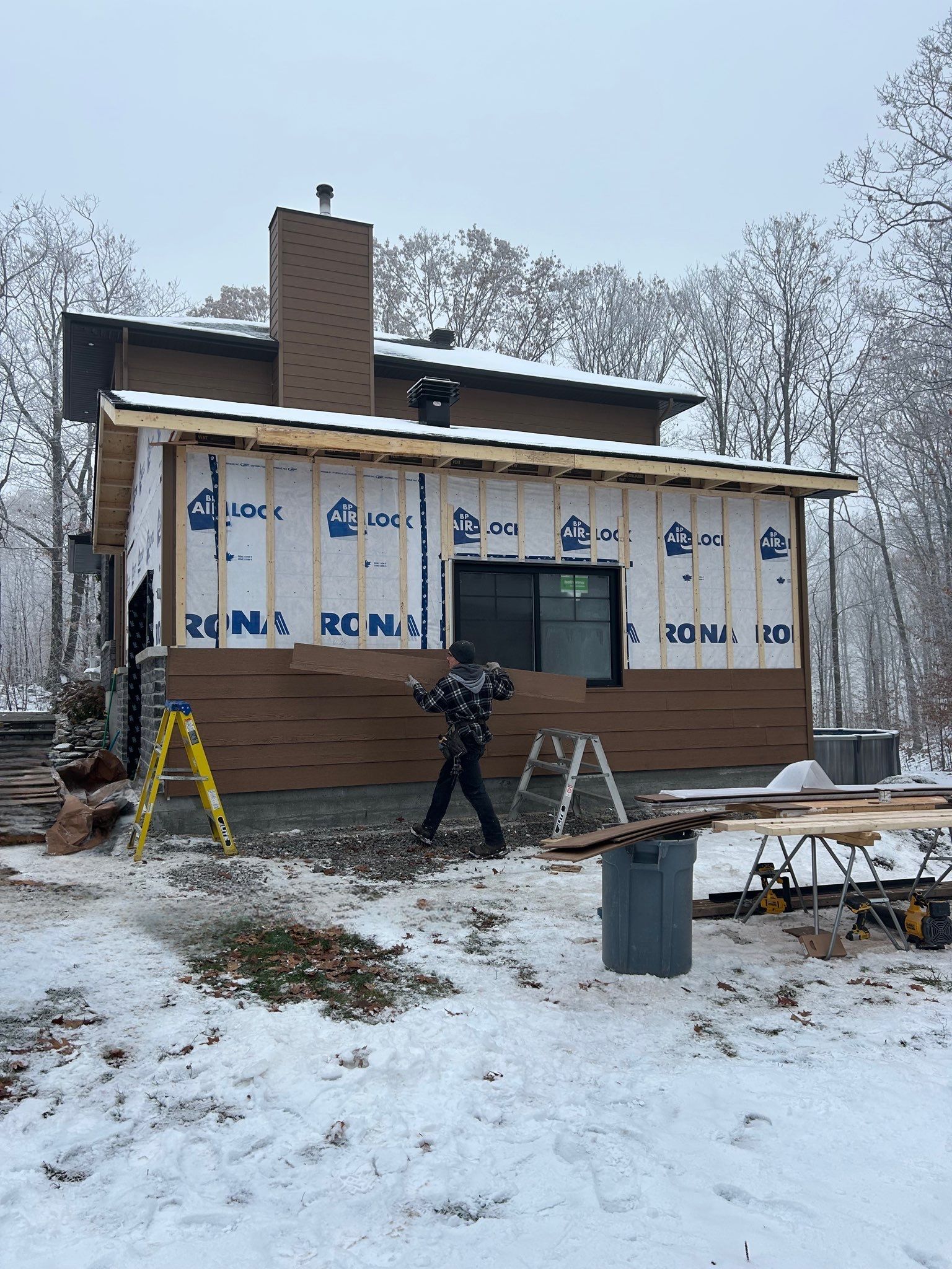 Maison enneigée en construction : des ouvriers installent le bardage et une cheminée sur la façade extérieure.