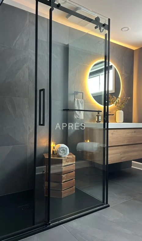 Salle de bains moderne avec portes de douche en verre noir, meuble-lavabo en bois et miroir rond lumineux.
