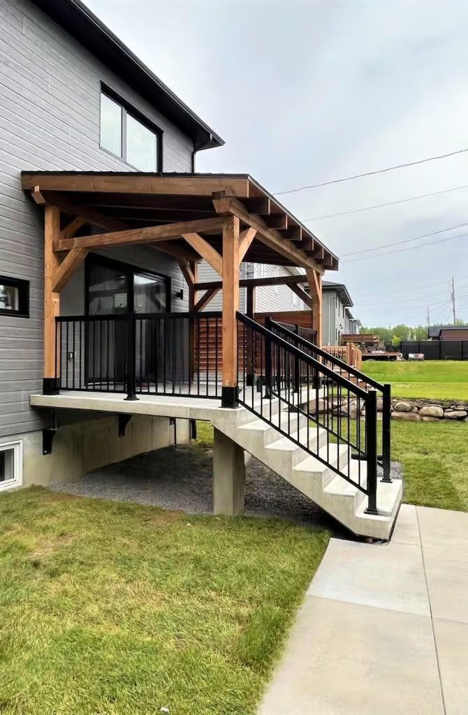 Extérieur de la maison avec porche en bois, balustrade noire et marches en béton menant à une cour gazonnée