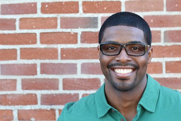 A man wearing glasses and a green shirt is smiling in front of a brick wall.