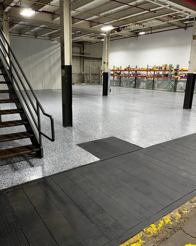 Empty industrial warehouse with black metal stairs, black columns, and a gray epoxy floor.