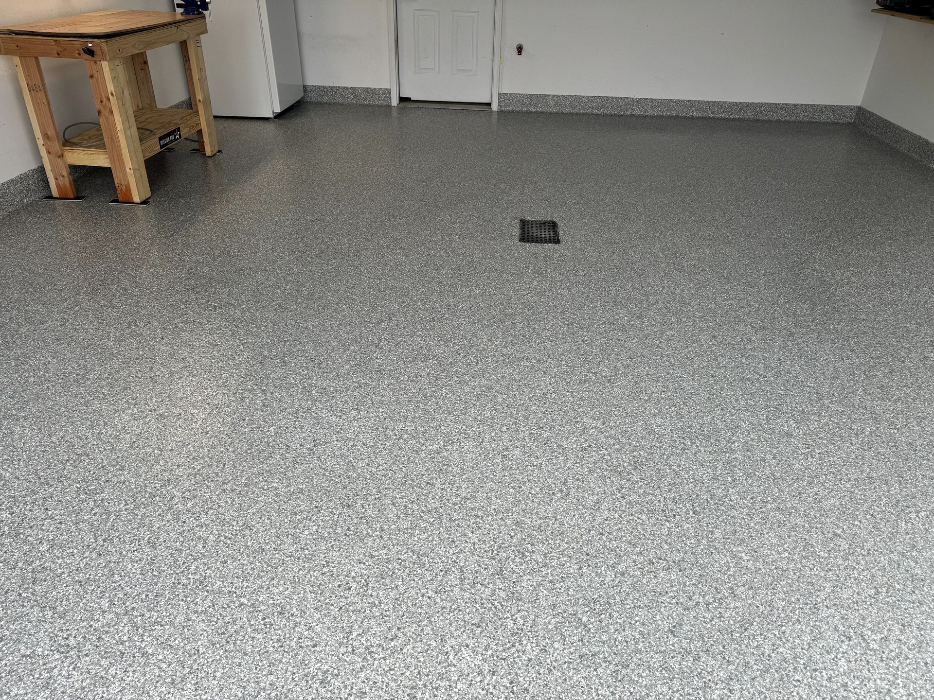 Empty gray speckled floor in a room, with a small black object near the center and a wooden table at left.