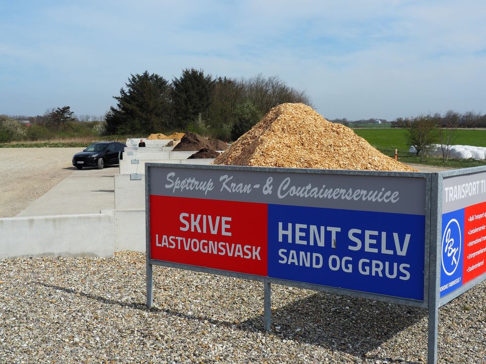 Skilt ved en affaldsplads med træflis og grus, ved siden af ​​en parkeret bil.