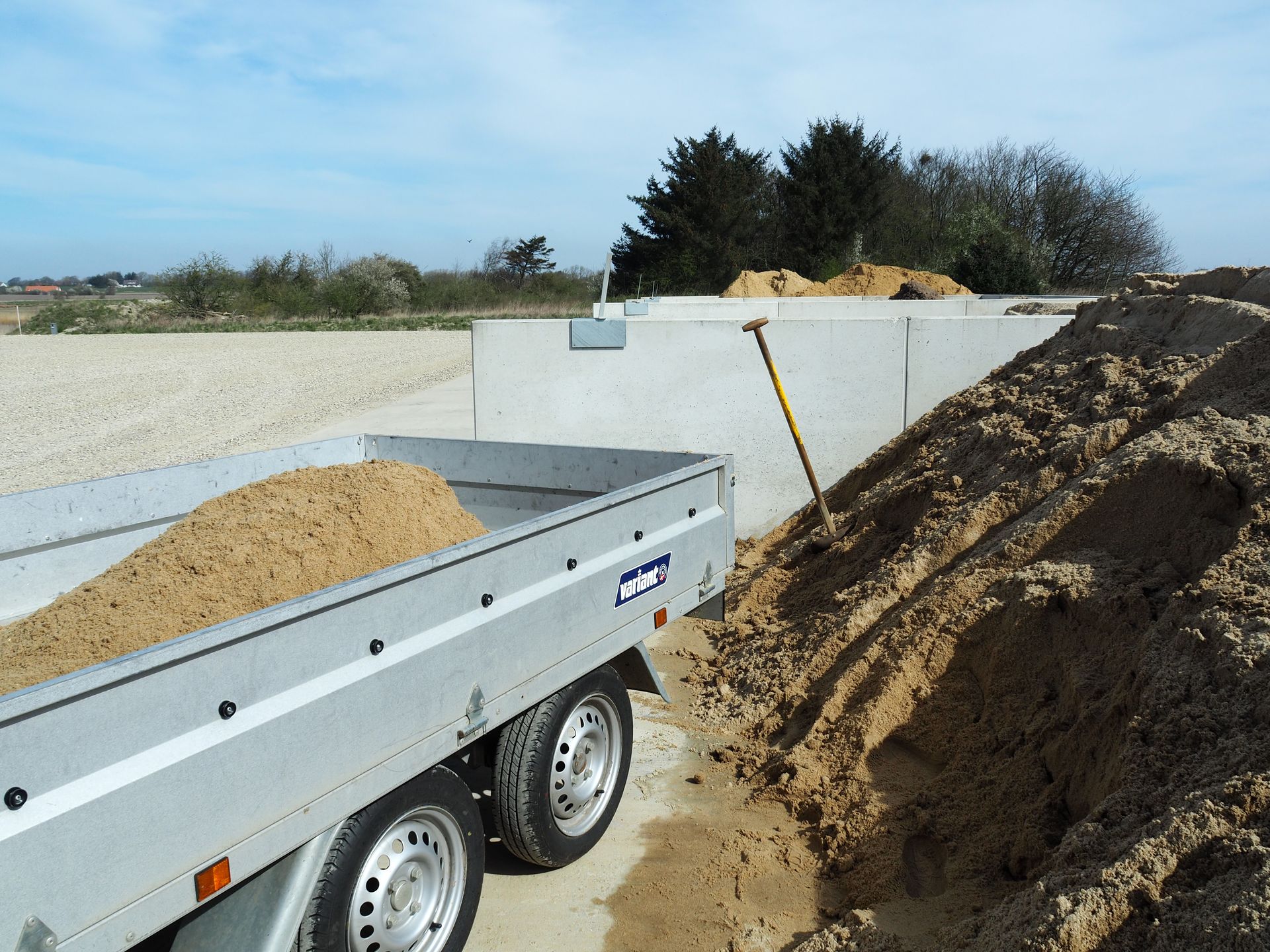 En sølvfarvet trailer ved siden af ​​en bunke sand med en betonvæg i baggrunden på en udendørs byggeplads.