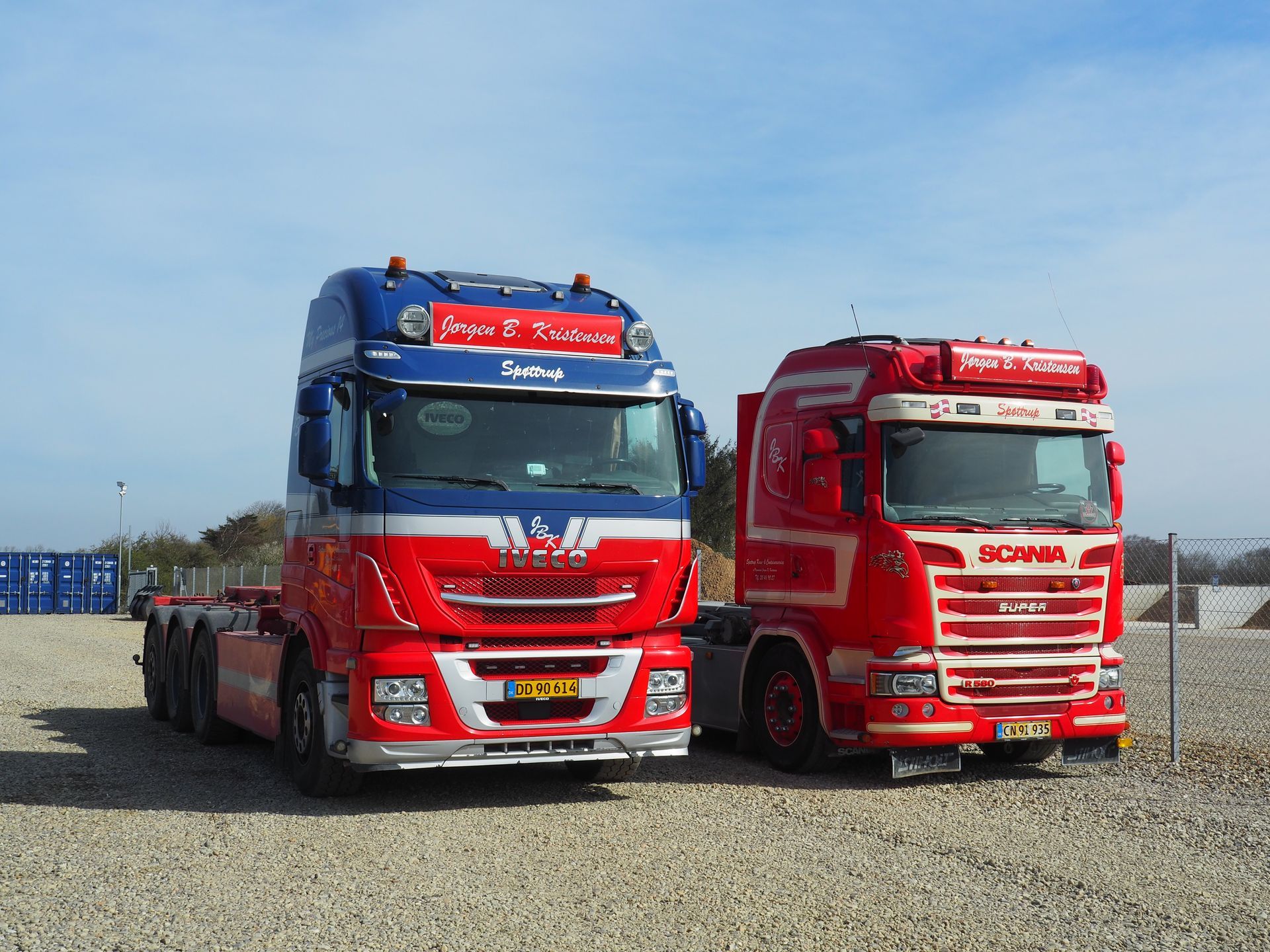 To farverige semitrucks parkeret side om side på en grusplads, den ene blå og rød, den anden rød med hvide detaljer.